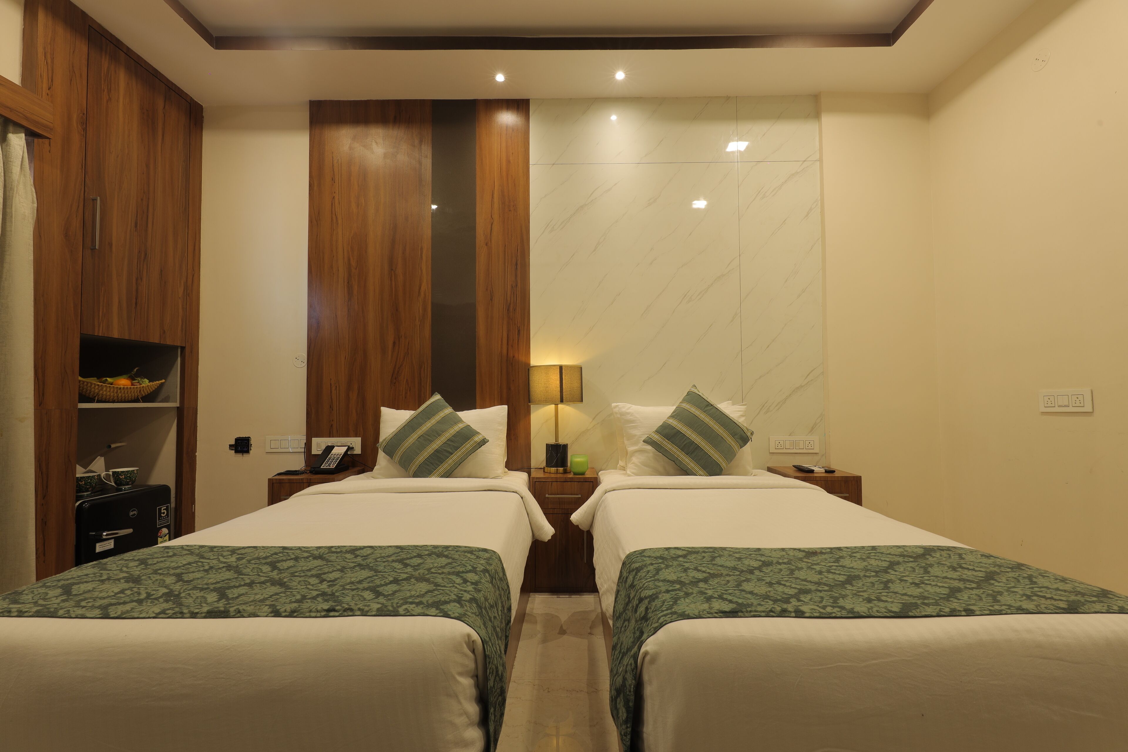 A hotel room with two beds, wooden wardrobe, marble wall, and a small shelf with a basket.