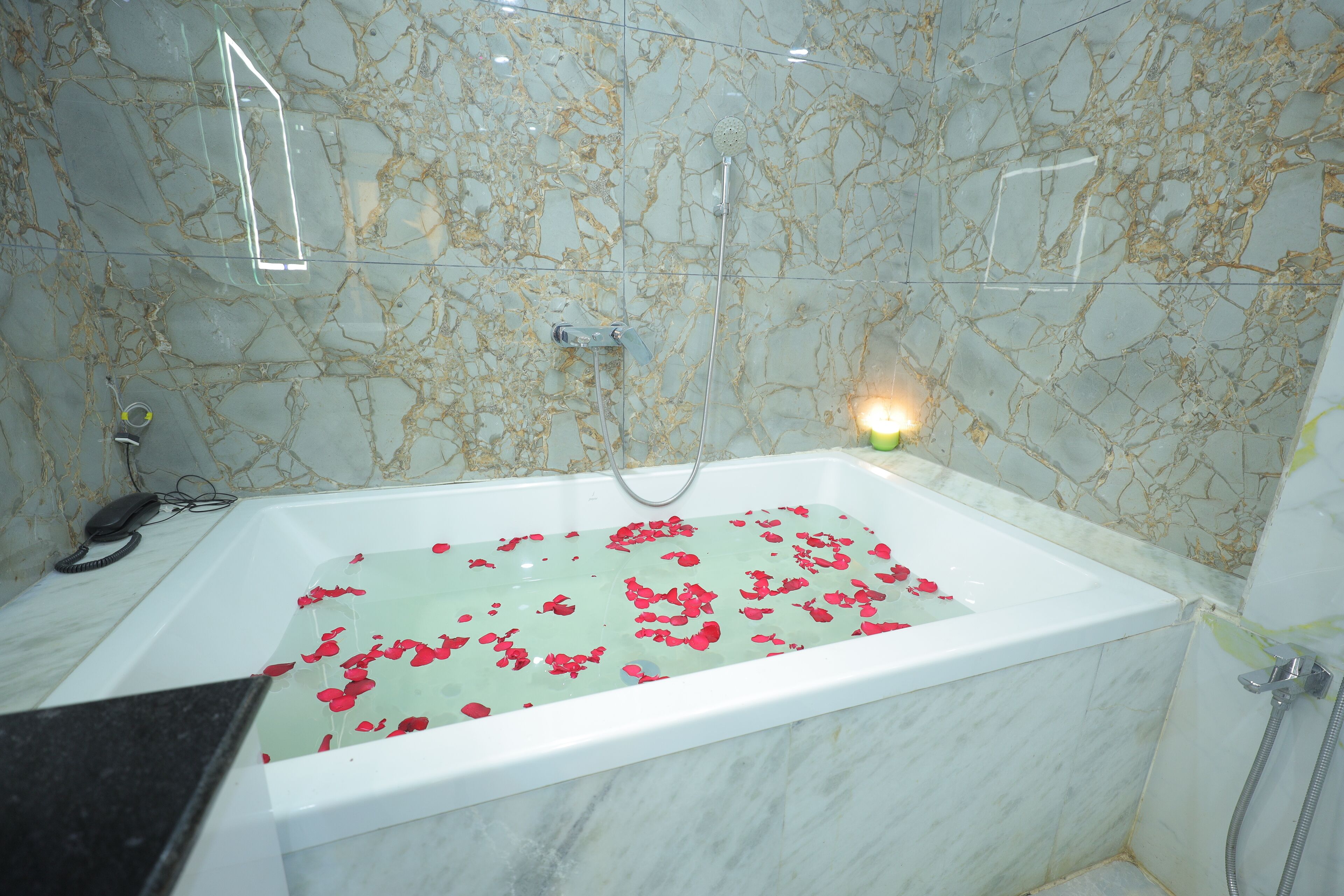A bathroom with a marble wall, a bathtub filled with water and rose petals, a handheld showerhead, and a small lit candle.