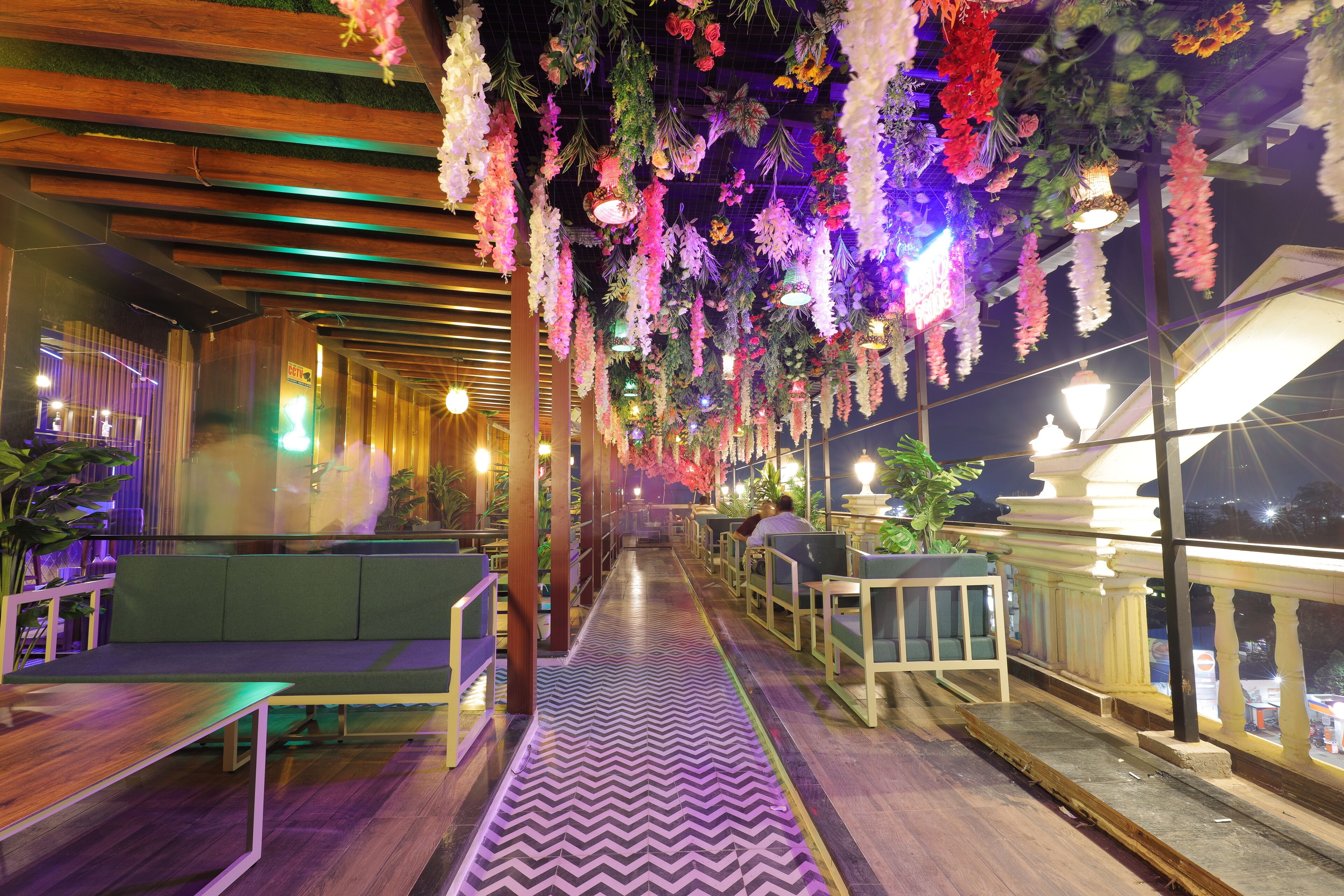 A restaurant with a long, narrow layout featuring a central walkway, seating areas on both sides, and hanging floral decorations.