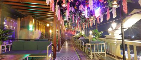 A restaurant with a long, narrow layout featuring a central walkway, seating areas on both sides, and hanging floral decorations.