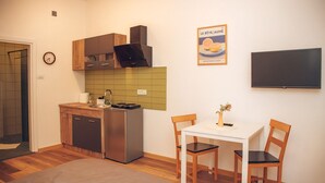 A compact kitchen with a refrigerator, stove, and wooden cabinets. There is a dining table with two chairs and a small vase. A flat-screen TV is mounted on the wall.