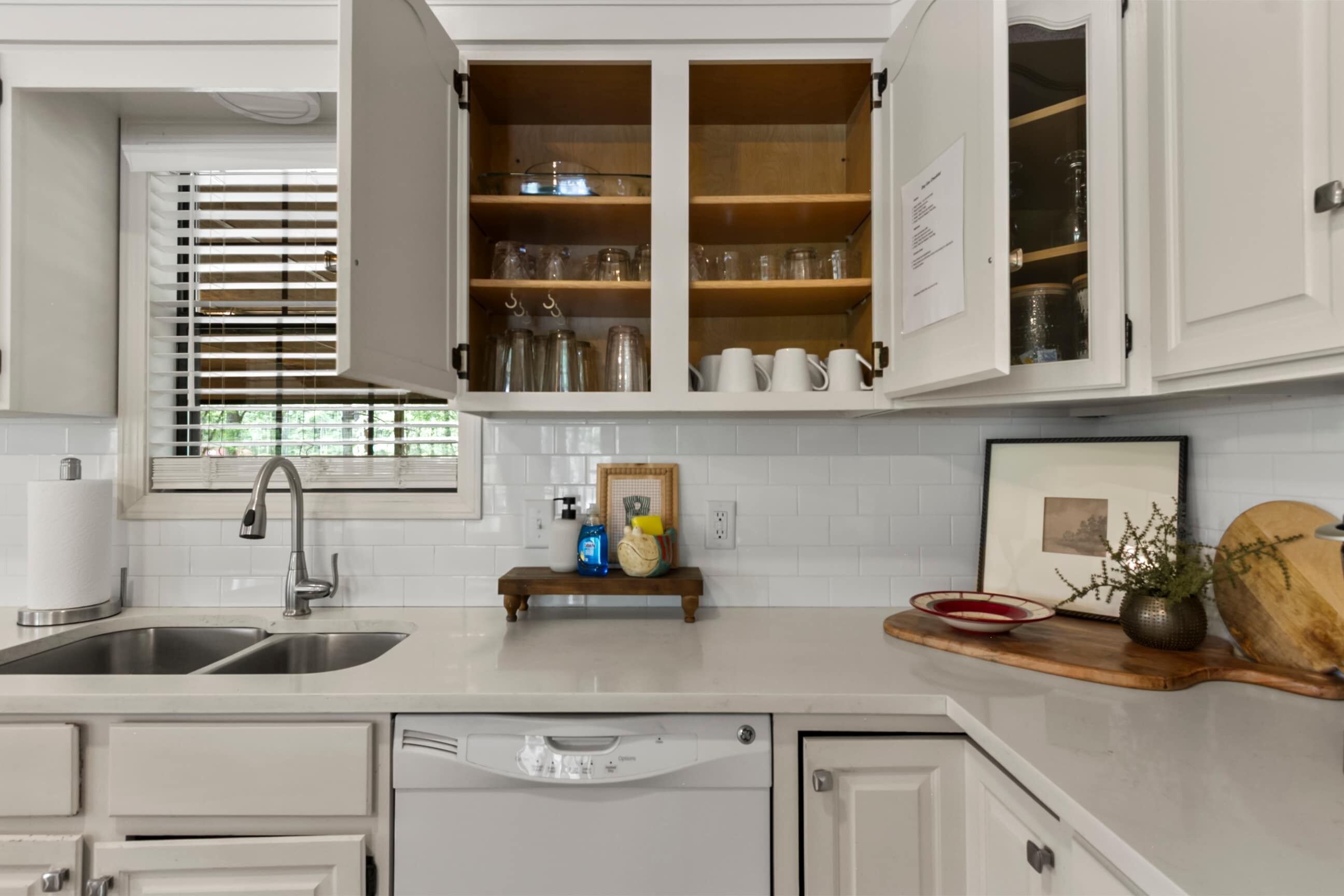 Una cocina con armarios blancos, lavavajillas, lavabo y ventana con persianas.