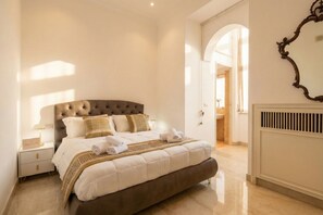 Une chambre bien éclairée, avec une tête de lit capitonnée, un lit soigneusement fait, une table de chevet avec une lampe et un radiateur au mur.