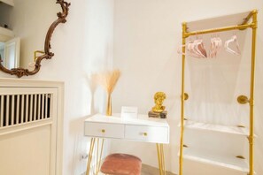 Uma toucador branca com detalhes dourados, um espelho com moldura ornamentada, uma estátua dourada e uma prateleira dourada com roupas penduradas.