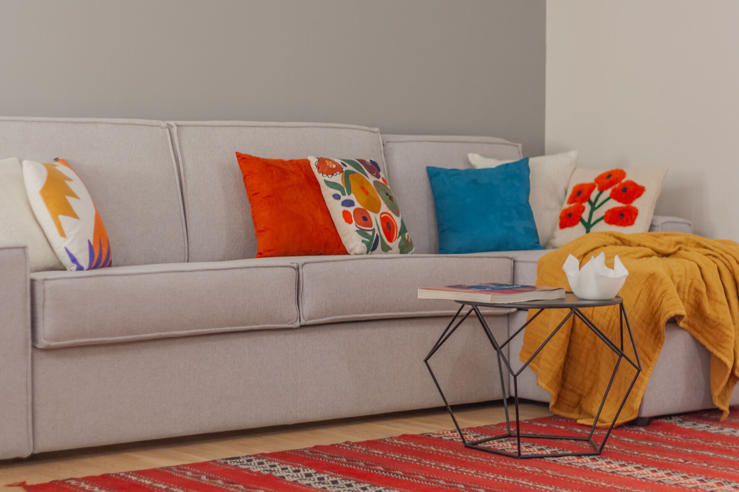 Un salon moderne avec un canapé gris, des coussins colorés, un tapis à motifs rouges et une petite table noire sur laquelle se trouvent un livre et un vase blanc.