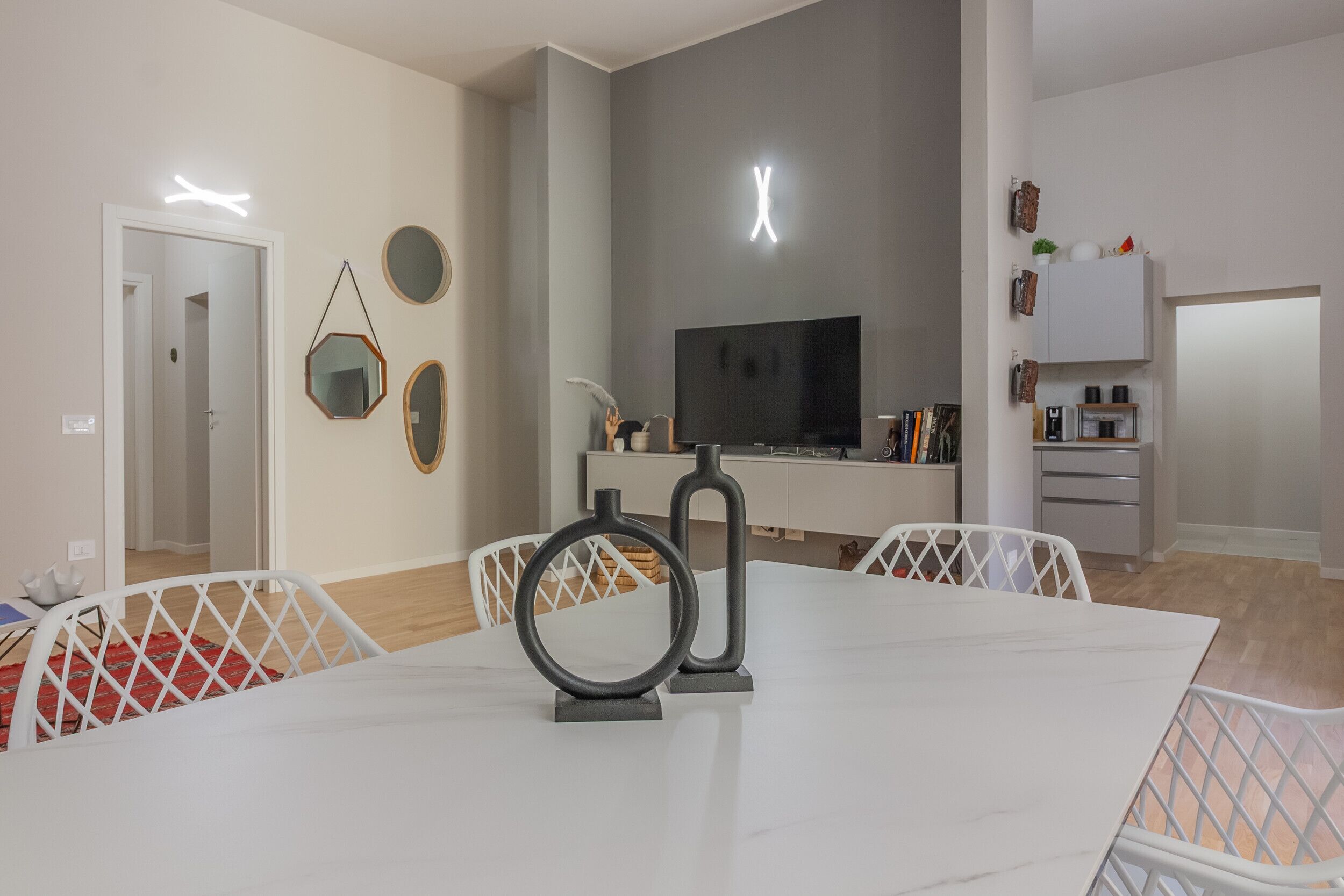 A modern living room with a white dining table, a flat-screen TV, and a kitchen area with cabinets and a microwave.