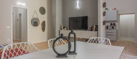 A modern living room with a white dining table, a flat-screen TV, and a kitchen area with cabinets and a microwave.