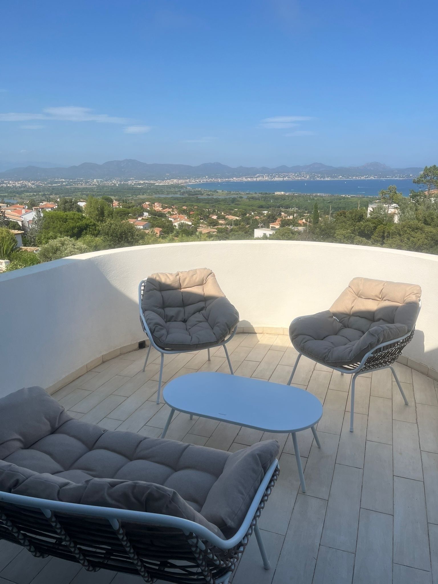 A terrace with a sofa, two chairs, and a small table. The terrace overlooks a coastal cityscape with mountains in the distance.