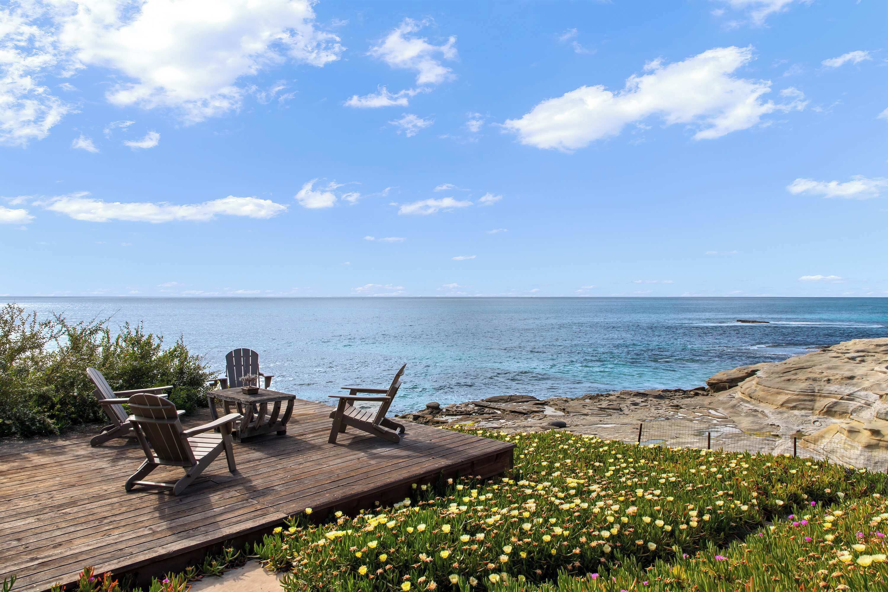 Una terraza de madera con sillas Adirondack y una mesa con vista a una costa rocosa y una vista despejada del océano.