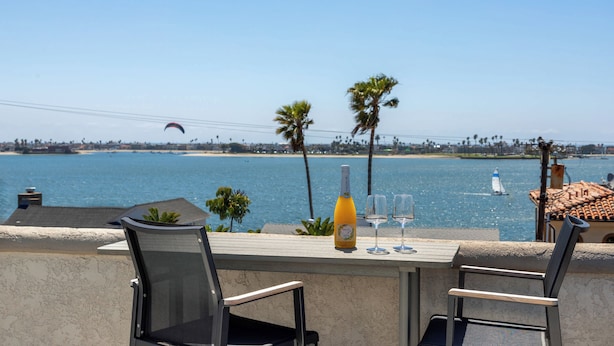 A terrace with a table, two chairs, a bottle of wine, and glasses, overlooking a beach with palm trees and a sailboat.