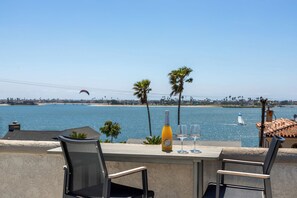 Une terrasse avec une table, deux chaises, une bouteille de vin et des verres, donnant sur une plage bordée de palmiers et où l’on aperçoit un voilier.