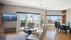 A spacious living room with a large TV, comfortable seating, a dining area, and a view of the sea through large windows.