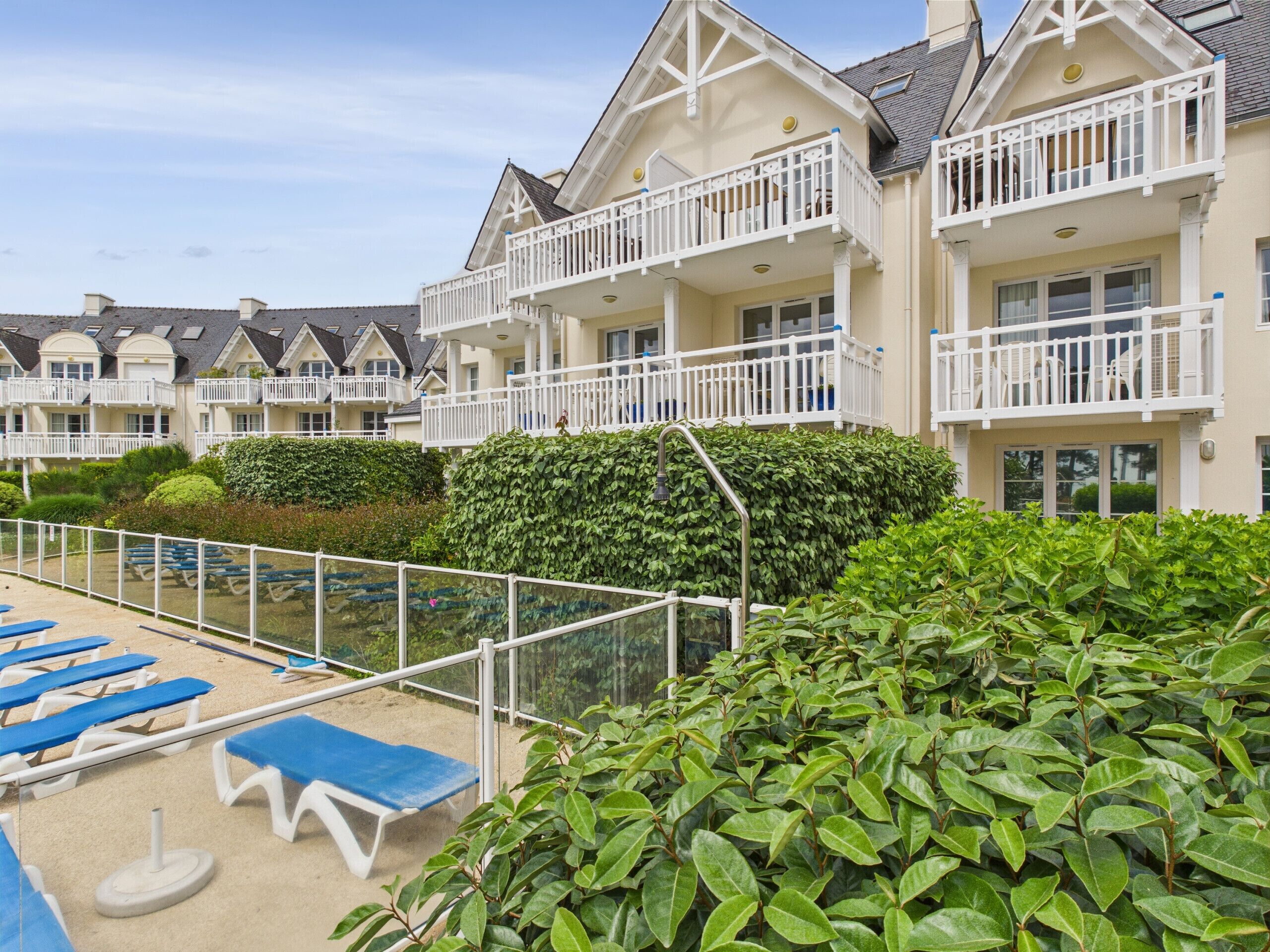 A row of seaside cottages with balconies, a sandy beach area with blue lounge chairs, and dense green shrubbery.