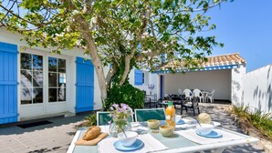 An outdoor dining area with a table set for two, surrounded by a white building with blue shutters, a tree, and a clear blue sky. - 500 m from the beach - House for 6 - Terrace (L'Epine)