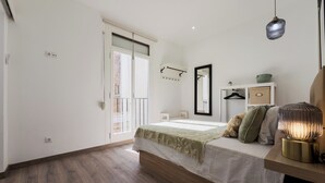 Un dormitorio con cama, mesita de noche, lámpara colgante, espejo y ventana con vistas a edificios.