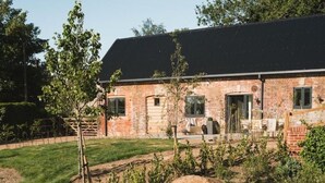 A brick house with a black roof, a front porch, and a small garden area with a tree and a rock. - Longstones Avebury – Ridgeway (Marlborough)