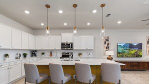 A modern kitchen with white cabinets, a central island with bar stools, stainless steel appliances, pendant lighting, and a large flat-screen TV on the wall.