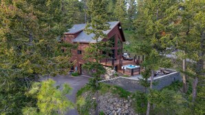A modern log cabin with a hot tub and outdoor seating area surrounded by dense forest.