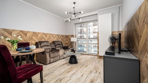 A modern living room with a sofa, a dining table with chairs, a standing fan, and a large window with a view of buildings. - Apartamenty Sun&Snow Platinum Towers (Warsaw)
