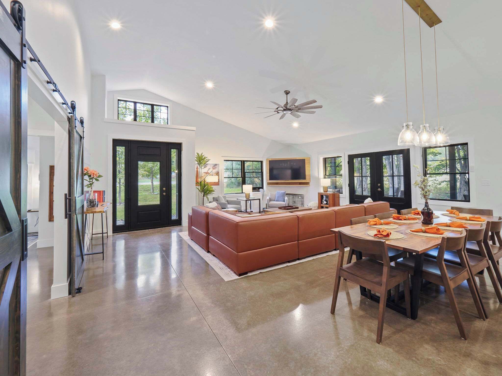 A spacious living area with a large sofa, dining table, and chairs. There is a TV mounted on the wall and a ceiling fan.