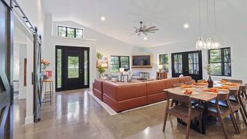 A spacious living area with a large sofa, dining table, and chairs. There is a TV mounted on the wall and a ceiling fan.