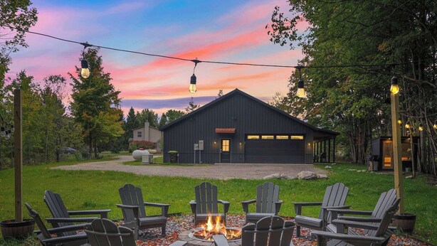 A rustic outdoor seating area with Adirondack chairs around a fire pit, a modern building in the background, and a string of lights above.