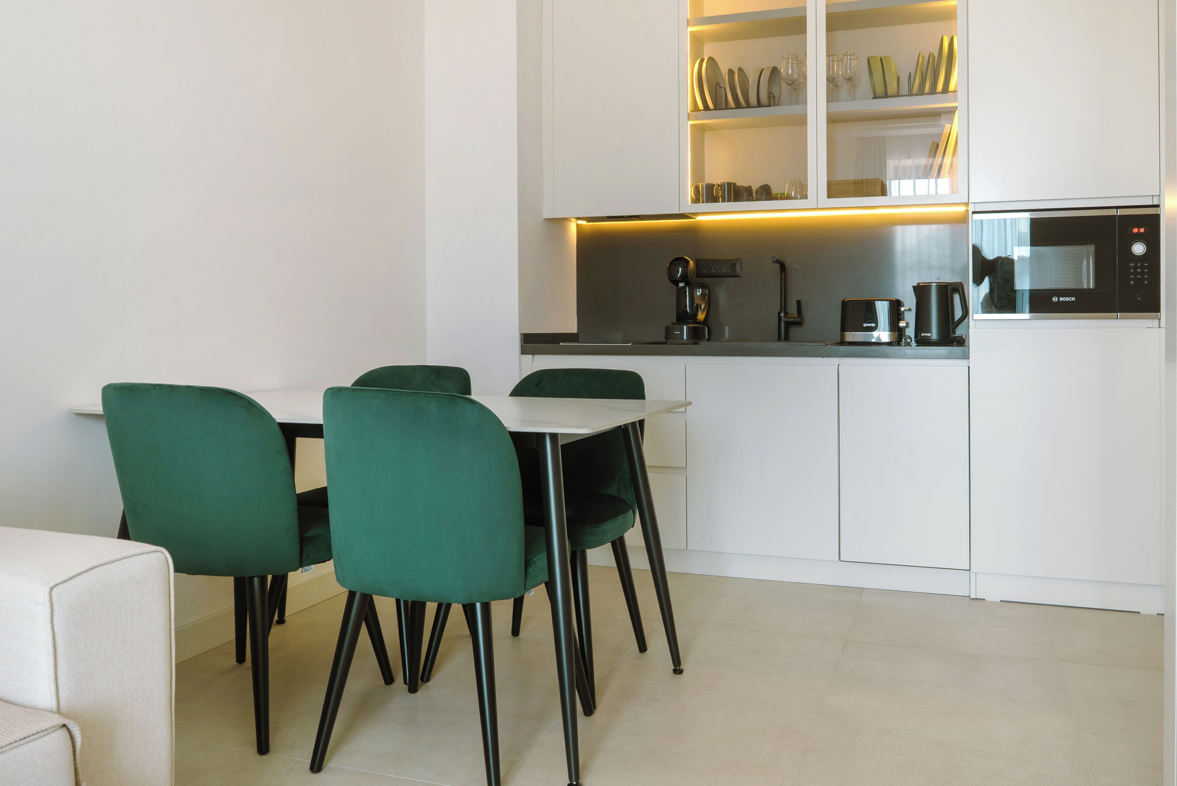 Una cocina moderna con armarios blancos, un microondas Bosch y una cafetera. Cuatro sillas verdes rodean una mesa blanca.
