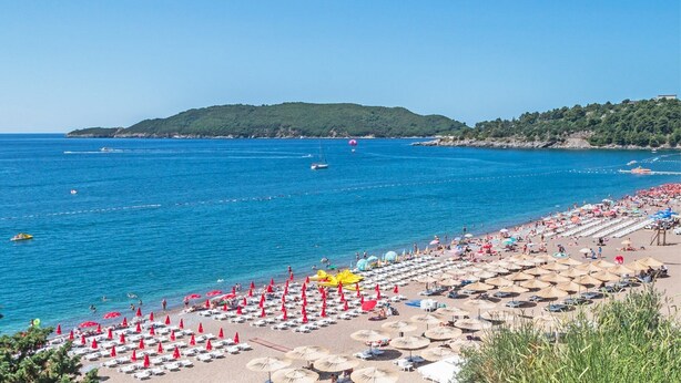 Une plage bordée de rangées de transats et de parasols, une mer d’un bleu limpide et, au loin, un paysage vallonné.