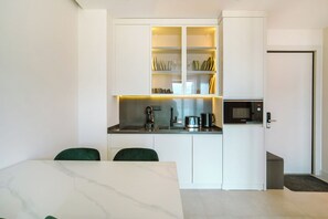 A compact kitchen with white cabinetry, a built-in microwave, and a coffee maker. There are two green chairs and a marble table in the adjacent dining area.