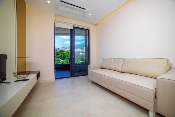 Ein modernes Wohnzimmer mit einem beigefarbenen Sofa, einem Fernseher und einer Glastür, die auf einen Balkon mit Stadtblick führt.