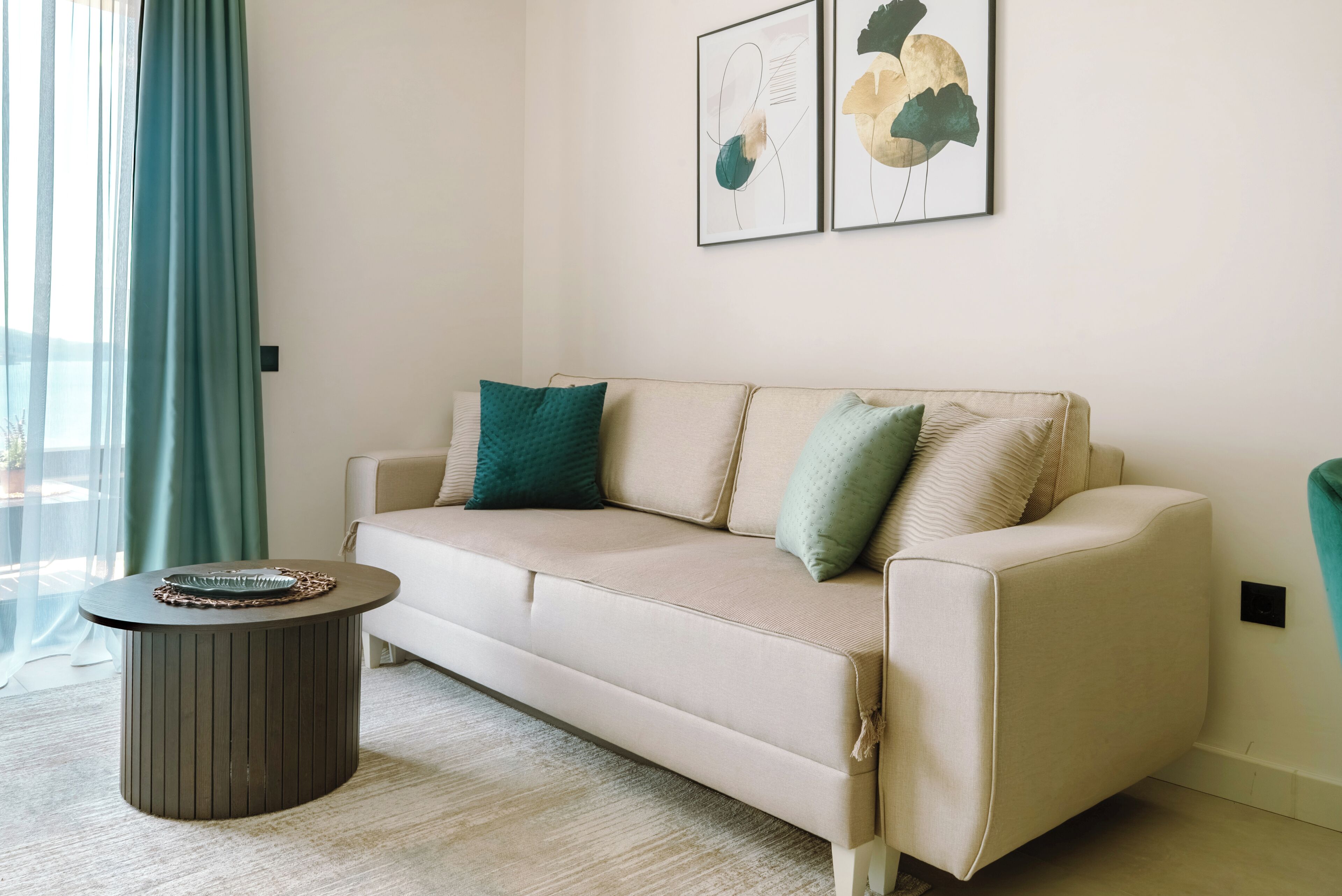Un salón con un sofá beige, dos almohadas, una mesa de centro redonda y arte abstracto en la pared.