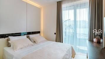 A hotel room with a large bed, a view of a cityscape through sheer curtains, and a bedside table with a vase of flowers.