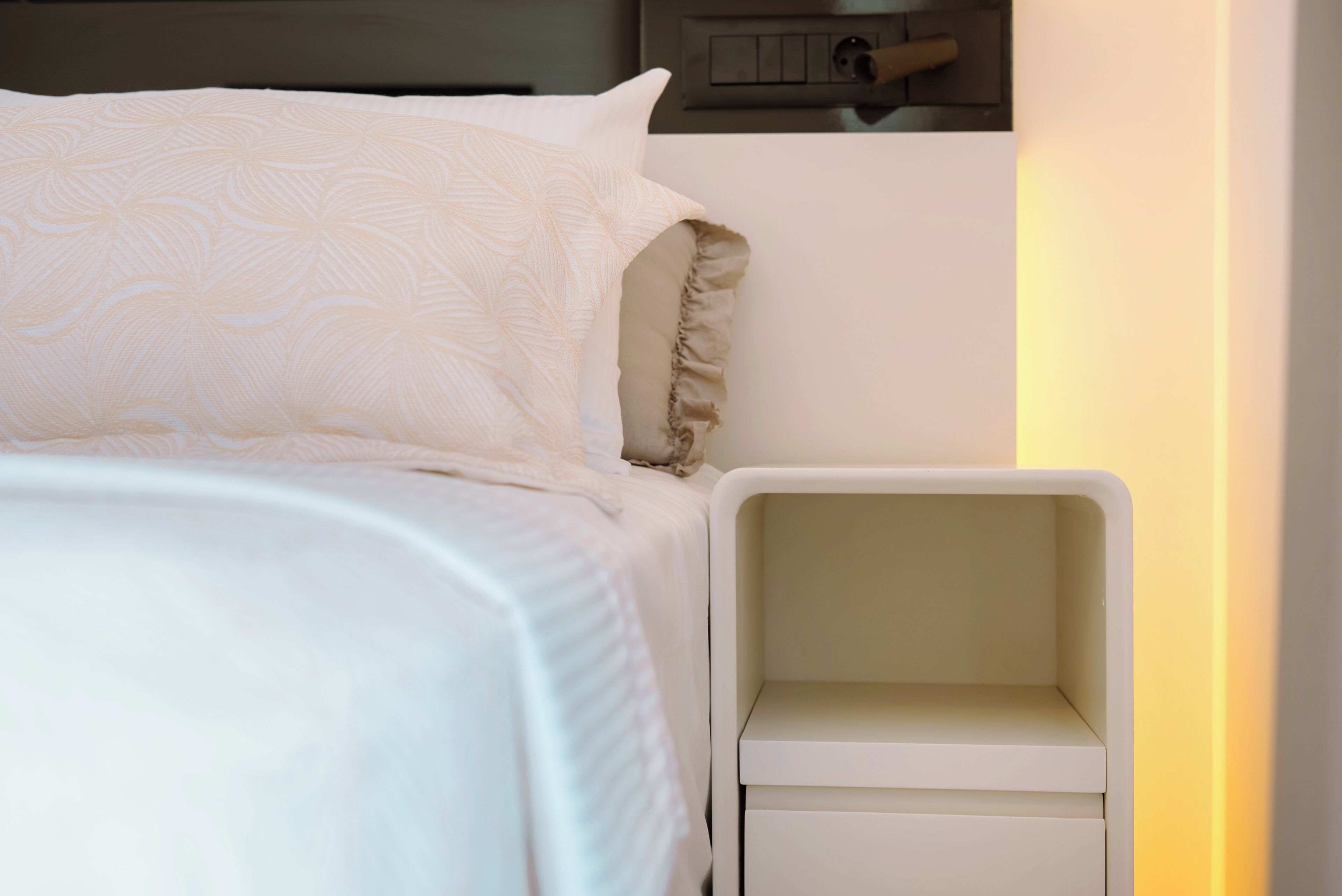 Una cama bien hecha con edredón blanco estampado y almohadas, una mesita de noche blanca con cajones, y un panel de interruptores fijado a la pared.