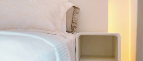 Una cama bien hecha con edredón blanco estampado y almohadas, una mesita de noche blanca con cajones, y un panel de interruptores fijado a la pared.