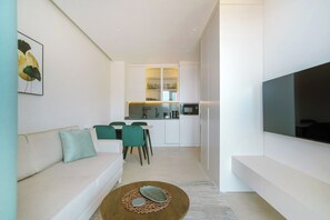A modern living room with a sofa, a coffee table, and a TV. A kitchen is visible in the background with cabinets and a dining area.