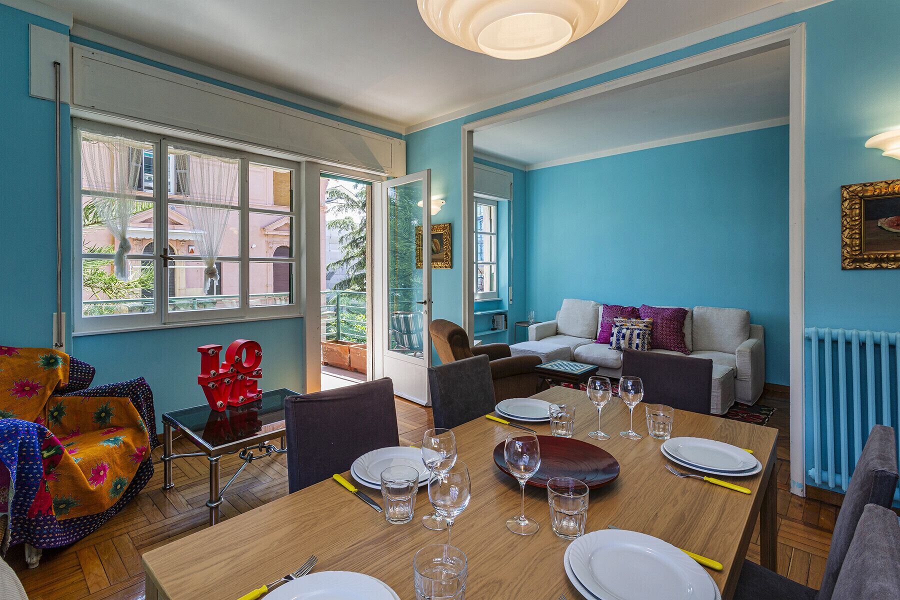 A dining area with a wooden table set for four, surrounded by chairs. There is a sofa and a small table with a decorative item in the background.