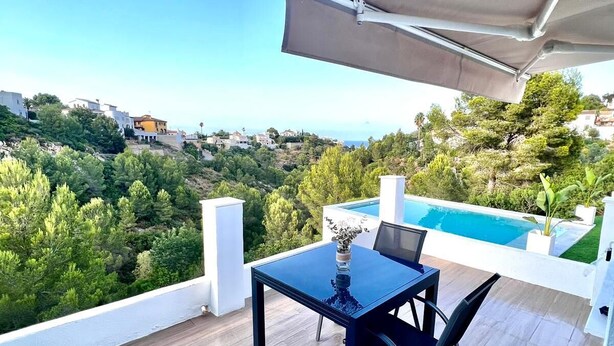 Un balcón con mesa y sillas, una piscina y vistas a una colina con casas.