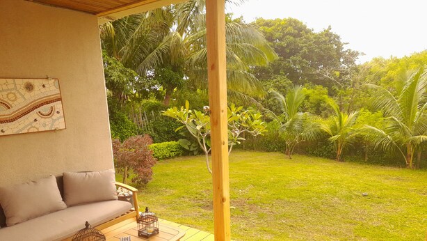Un espacio de estar al aire libre techado con terraza de madera, un sofá acolchado y vistas a un jardín exuberante con palmeras y césped.