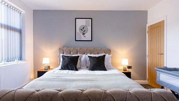 A neatly arranged bedroom with a tufted headboard, two bedside lamps, a framed artwork on the wall, and a window with curtains. - EXCLUSIVE MODERN NEWLY REFURBISHED HOME / CENTRAL LOCATION (Stoke-on-Trent)