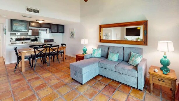 Una sala de estar con un sofá modular, una mesa de comedor con sillas, una cocina al fondo y un ventilador de techo.