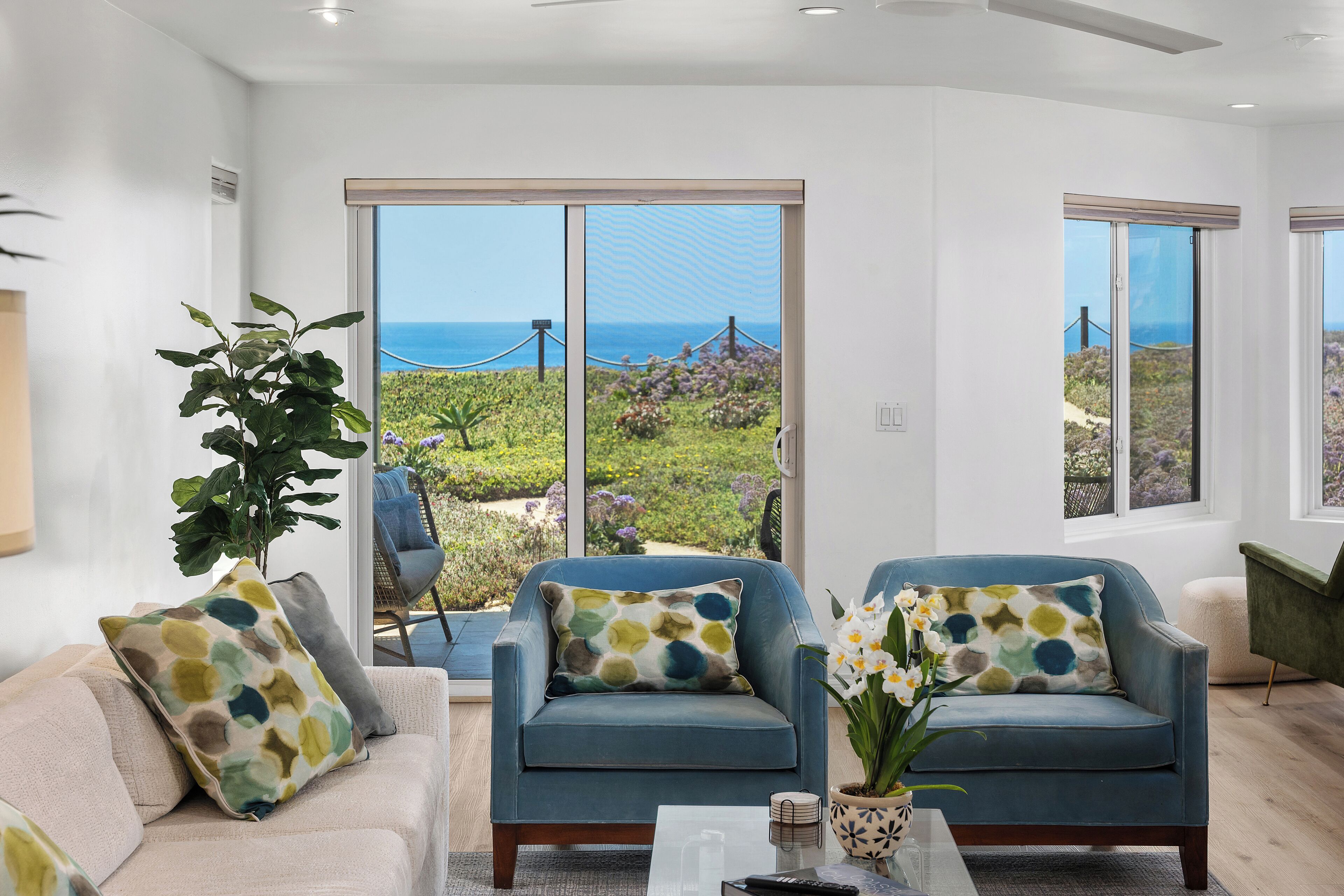 A modern living room with a large sliding glass door leading to a garden with a view of the sea. There are two blue armchairs with patterned cushions, a beige sofa, and a coffee table with decorative items.