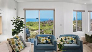 A modern living room with a large sliding glass door leading to a garden with a view of the sea. There are two blue armchairs with patterned cushions, a beige sofa, and a coffee table with decorative items. - Surf Song Symphony - Luxury Oceanfront Condo with Panoramic Views (Solana Beach)