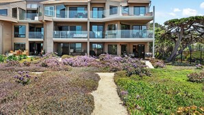 A multi-story residential building with balconies, a garden with purple flowers, and a clear sky. - Surf Song Symphony - Luxury Oceanfront Condo with Panoramic Views (Solana Beach)