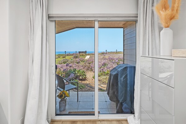 Vista a través de un ventanal corredizo a un patio exterior con jardín, una silla y una mesa.