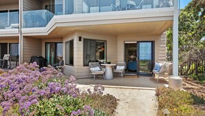 A modern building with a covered outdoor seating area, large windows, and a garden with purple flowers. - Surf Song Symphony - Luxury Oceanfront Condo with Panoramic Views (Solana Beach)