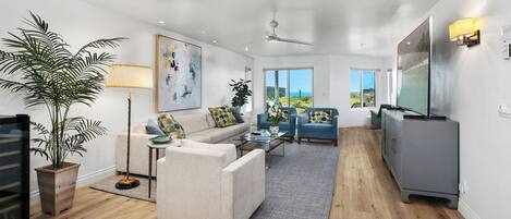 Una sala de estar moderna con un sofá, sillones, una mesa de centro y un televisor. Se aprecia una planta grande, un cuadro y vistas al exterior.