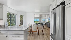 A modern kitchen with white cabinetry, a large stainless steel refrigerator, and a dining area with wooden chairs and a glass table. - Surf Song Symphony - Luxury Oceanfront Condo with Panoramic Views (Solana Beach)