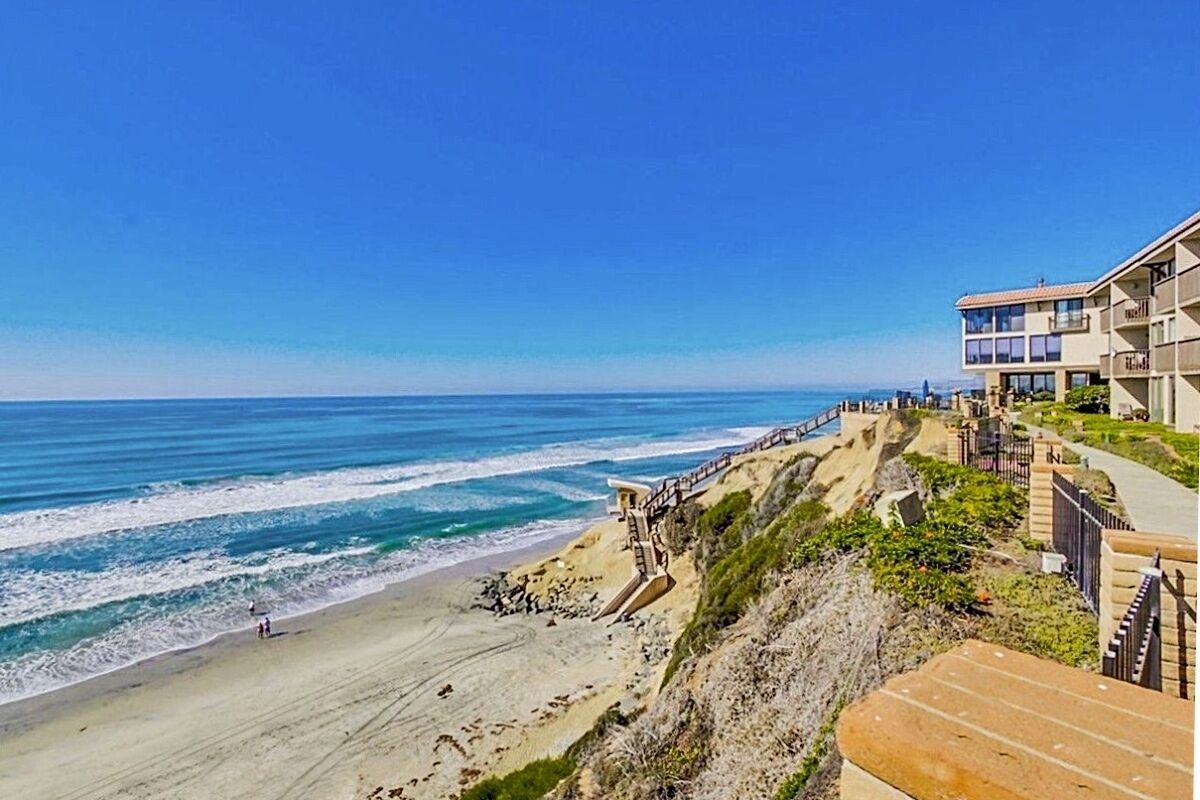 Ein Hotel an der Küste mit Blick auf den Strand, einer Treppe, die zum Strand hinunterführt, und einem klaren, blauen Himmel.