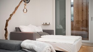 Quarto moderno com sofá cinza, cama branca, piso de madeira, porta de vidro e prateleira na parede com uma planta.
