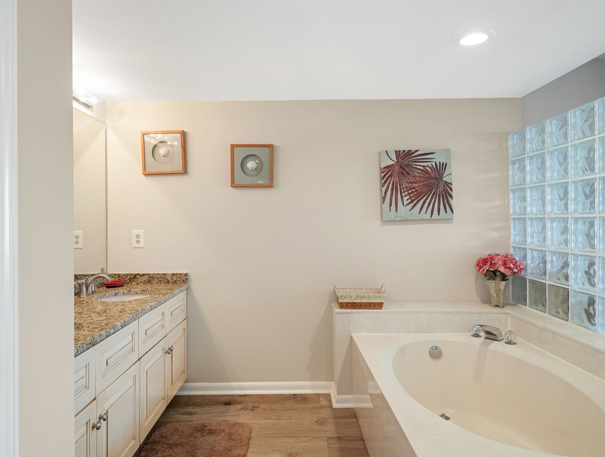 A bathroom with a white bathtub, a vanity with a granite countertop, and a mirror. There are two framed pictures on the wall and a vase with flowers on the vanity.